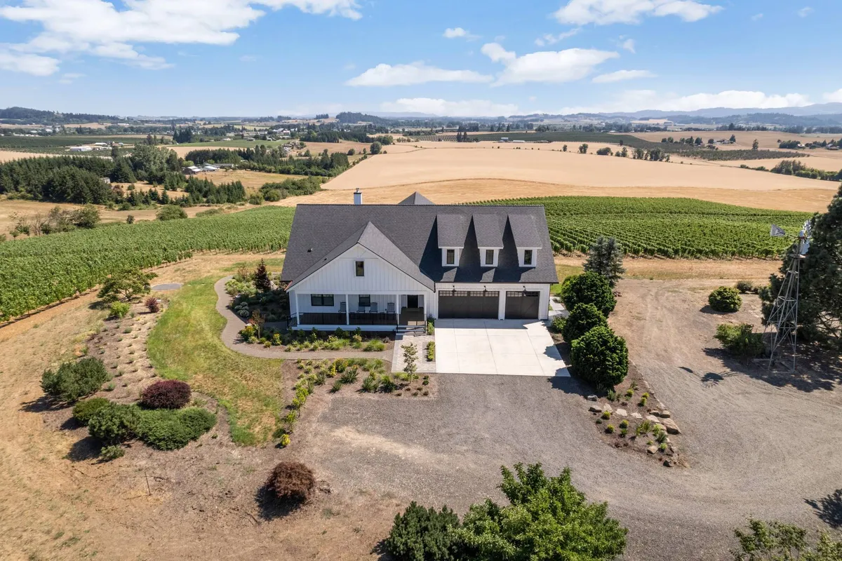 A sweeping exterior shot of a modern farmhouse surrounded by a landscaped garden, lush vineyards, and open fields. The scene captures the natural beauty and tranquility of the property, with plenty of space for outdoor gatherings and exploration.