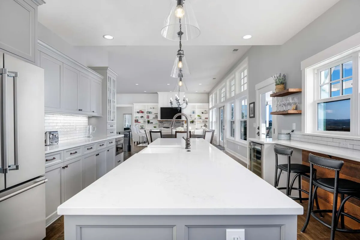 A spacious chef’s kitchen featuring a large kitchen island with sleek stone countertops, stainless steel appliances, open shelving for display, and a glamorous chandelier overhead. The ample countertop space and open layout make it ideal for both cooking and gathering.