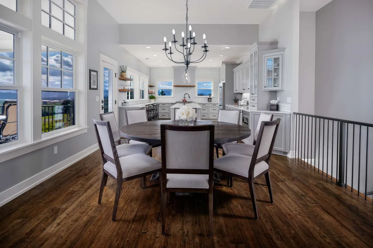 A round wooden dining table sits beneath a stylish chandelier, surrounded by comfortable chairs. Floor-to-ceiling windows on one side provide stunning views of the hills and vineyards, filling the space with warm natural light.