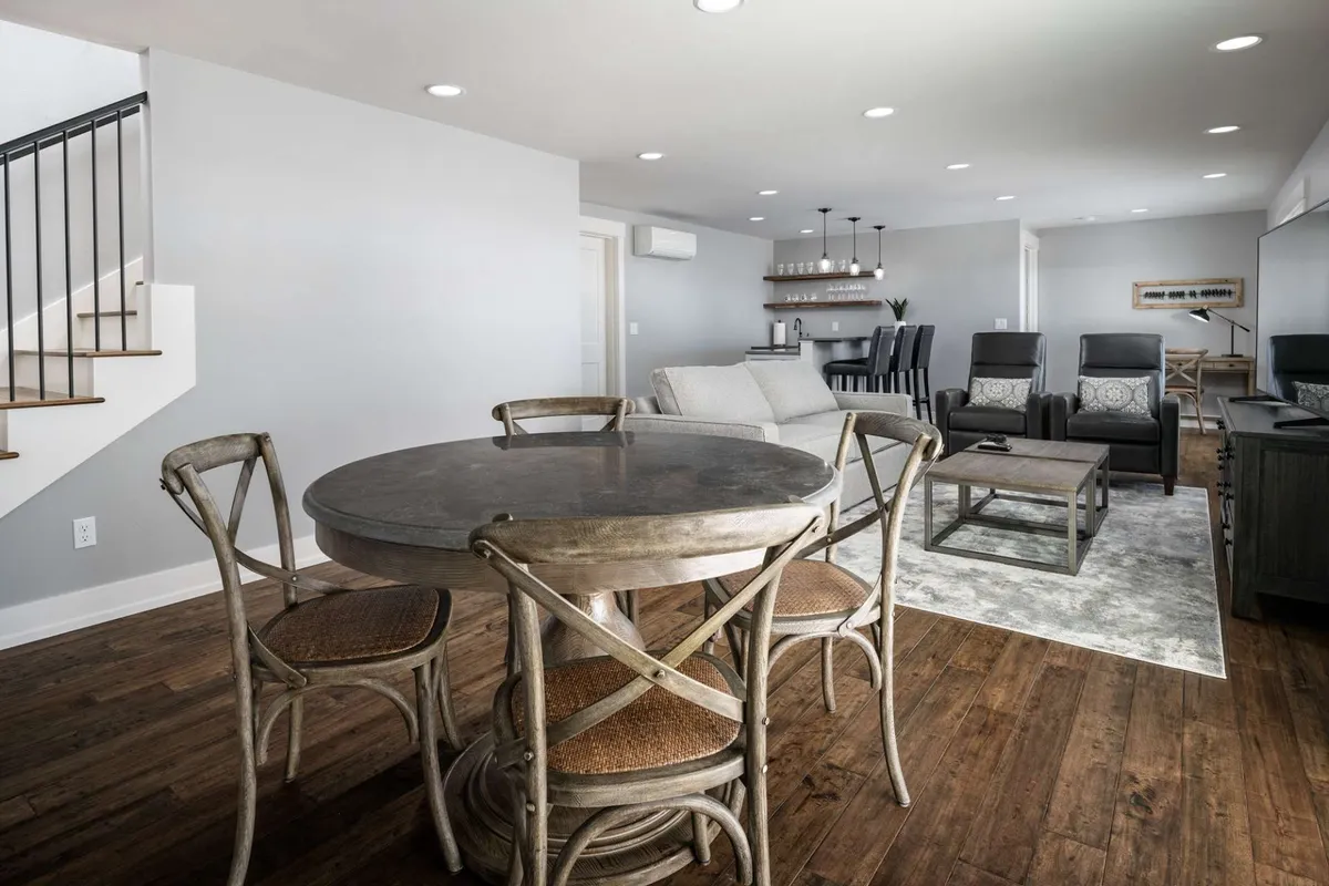 An open living and dining area with cozy, stylish seating arranged for conversation. The round dining table and a modern bar area are visible, set against a backdrop of large windows letting in natural light.