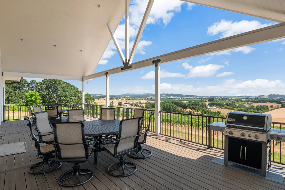 An elevated outdoor deck featuring a spacious dining table set for a meal, a grill nearby, and panoramic views of Oregon wine country hills. The scene captures a social, relaxed moment that suggests sunset dining and leisurely gatherings, with the horizon glowing in the background.