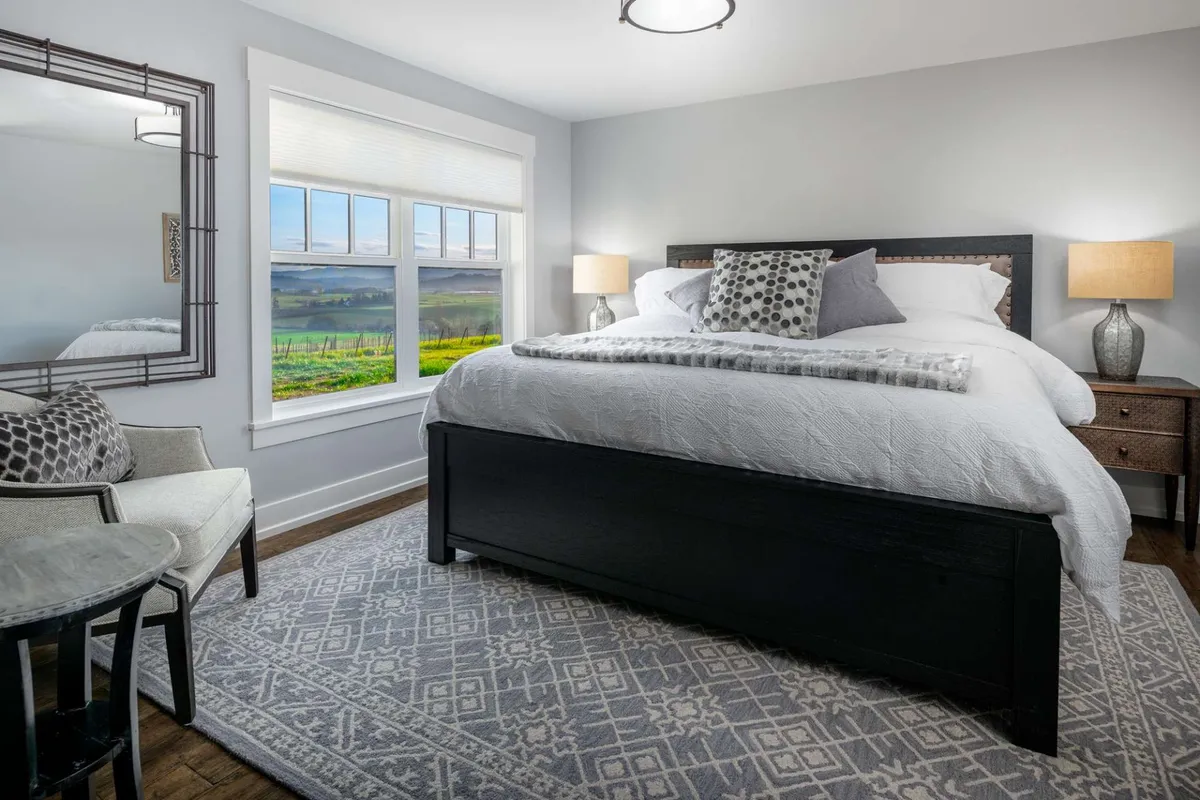 A spacious king-sized bed adorned with plush decorative pillows sits in a calming bedroom, flanked by side tables and lamps. The mirrored wall décor adds depth, while large windows frame picturesque vineyard views, flooding the room with natural light. The inviting setup emphasizes relaxation and luxury.