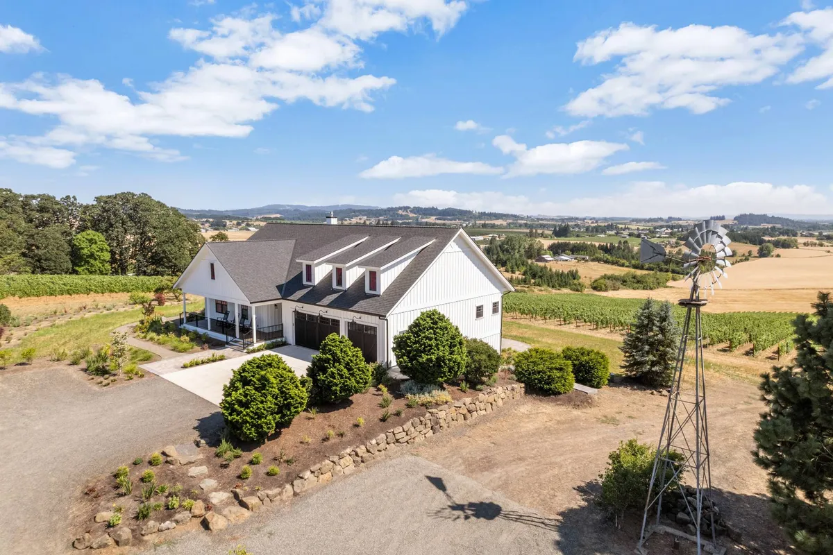 The house is nestled among lush vineyards, with a windmill and mature trees enhancing the sense of place. The setting is warm and inviting, suggesting opportunities for wine tasting, outdoor dining, and relaxation against the backdrop of spectacular vineyard sunsets.