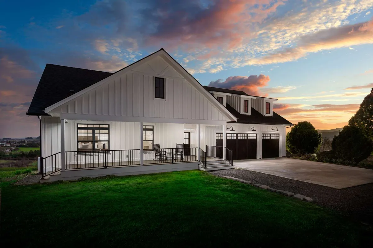 A welcoming white house is seen with a wide, inviting porch furnished with comfortable seating. The landscaped yard and garage doors suggest plenty of space for outdoor gatherings and relaxing in the fresh air. The setting sun casts a warm glow over the home, enhancing the sense of serenity and togetherness.