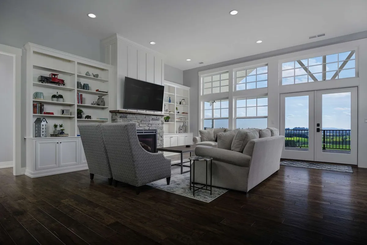 The image showcases a spacious and elegant living room with large windows framing sweeping vineyard views, a cozy fireplace as the focal point, plush seating arranged for conversation or relaxation, and built-in bookshelves. The soft natural light and inviting atmosphere set the stage for memorable gatherings, peaceful reading sessions, and enjoying sunsets over the Willamette Valley.