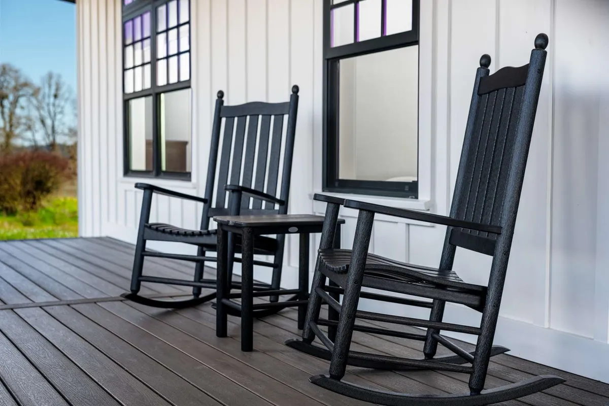A cozy outdoor porch scene with classic rocking chairs and a small side table, set against windows that reflect the peaceful surroundings. The atmosphere is calm and invites guests to unwind while enjoying the view or spending quiet moments in conversation.